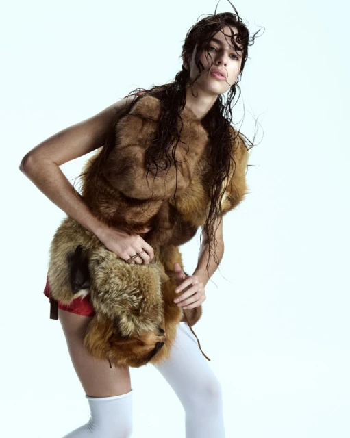 Young woman poses in a fur vest with white thigh-high socks in a studio setting. Fashion shot with wet hair and a white background.
