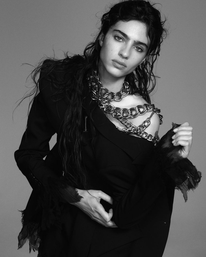 Monochrome portrait of a model with dark hair and a chain necklace, wearing a deconstructed black blazer in a studio setting.