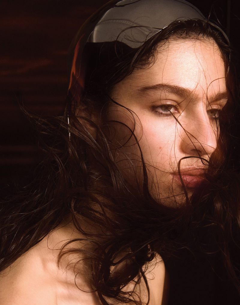 Close-up studio portrait of a woman with wet dark hair, glossy makeup, and a neutral expression, against a dark brown background.