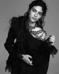 Model Loli Bahia wearing a black jacket and heavy silver chain necklace against a grey background