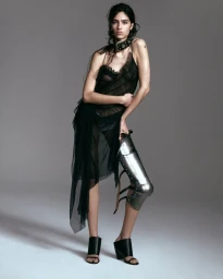 Model Loli Bahia wearing a sheer black dress and metallic leg armor against a grey studio backdrop.