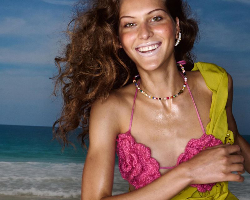 Woman in a pink bikini top and yellow cover-up poses at the beach with blue skies and ocean visible in the background on a sunny day.