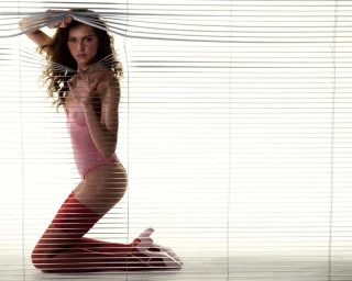Model Angelina Kendall peeking through window blinds wearing a pink bodysuit and patterned red stockings for MARFA.