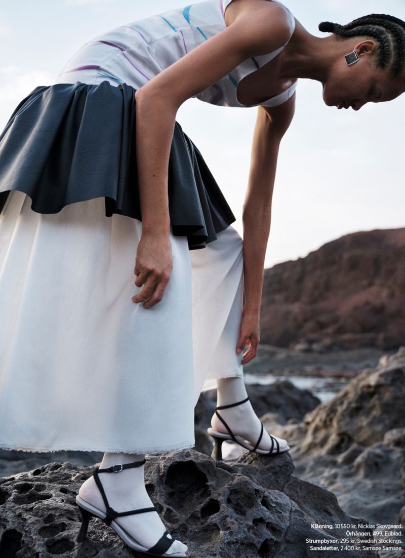 A slender woman in a layered dress and heels poses on a rocky surface, with a backdrop of blurred land under a bright sky.