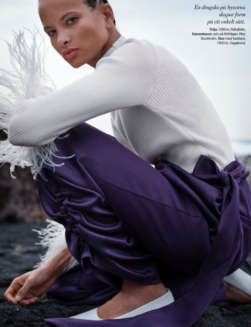 Fashion editorial shot of a woman in a white sweater and purple pants squatting on a rocky surface, showcasing clothing and style.