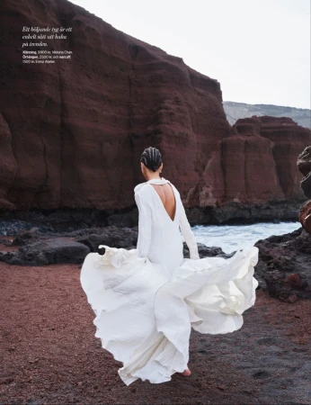 Woman in flowing white dress walks on red sand beach near ocean and cliffs, Santorini fashion shoot, travel editorial, lifestyle