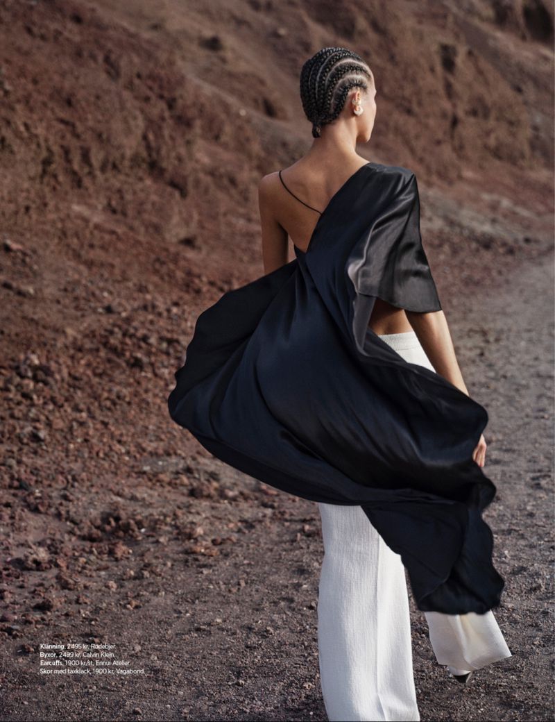 Adult female fashion model with braided hair poses in white pants and a draped black shawl in a rocky desert landscape with brown soil.