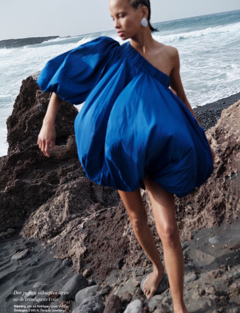 Fashion shot of a woman in an oversized cobalt dress posing on a rocky beach with the ocean behind her, earrings, and bare feet.