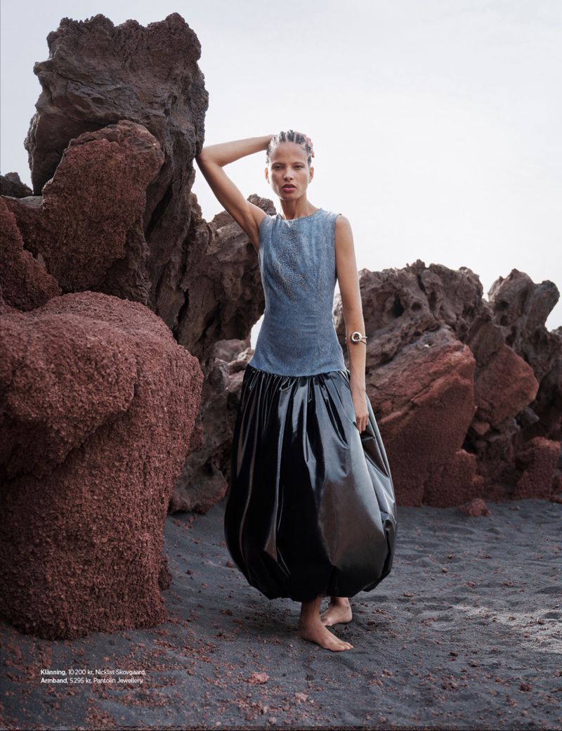 Fashion shot of a woman in a sleeveless top and oversized balloon skirt, posing barefoot on black sand with rocks in the background.