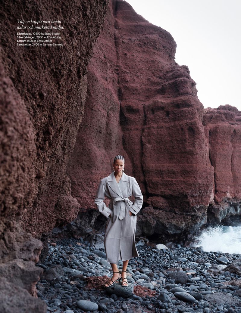 Fashion shot of a woman in a trench coat standing on a rocky beach, with red cliffs in the background and ocean waves visible.