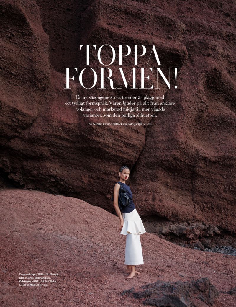 Fashion editorial shot of a woman in a dark top and light tiered skirt posing against a textured red rock formation outdoors.
