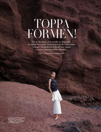 Fashion editorial shot of a woman in a dark top and light tiered skirt posing against a textured red rock formation outdoors.