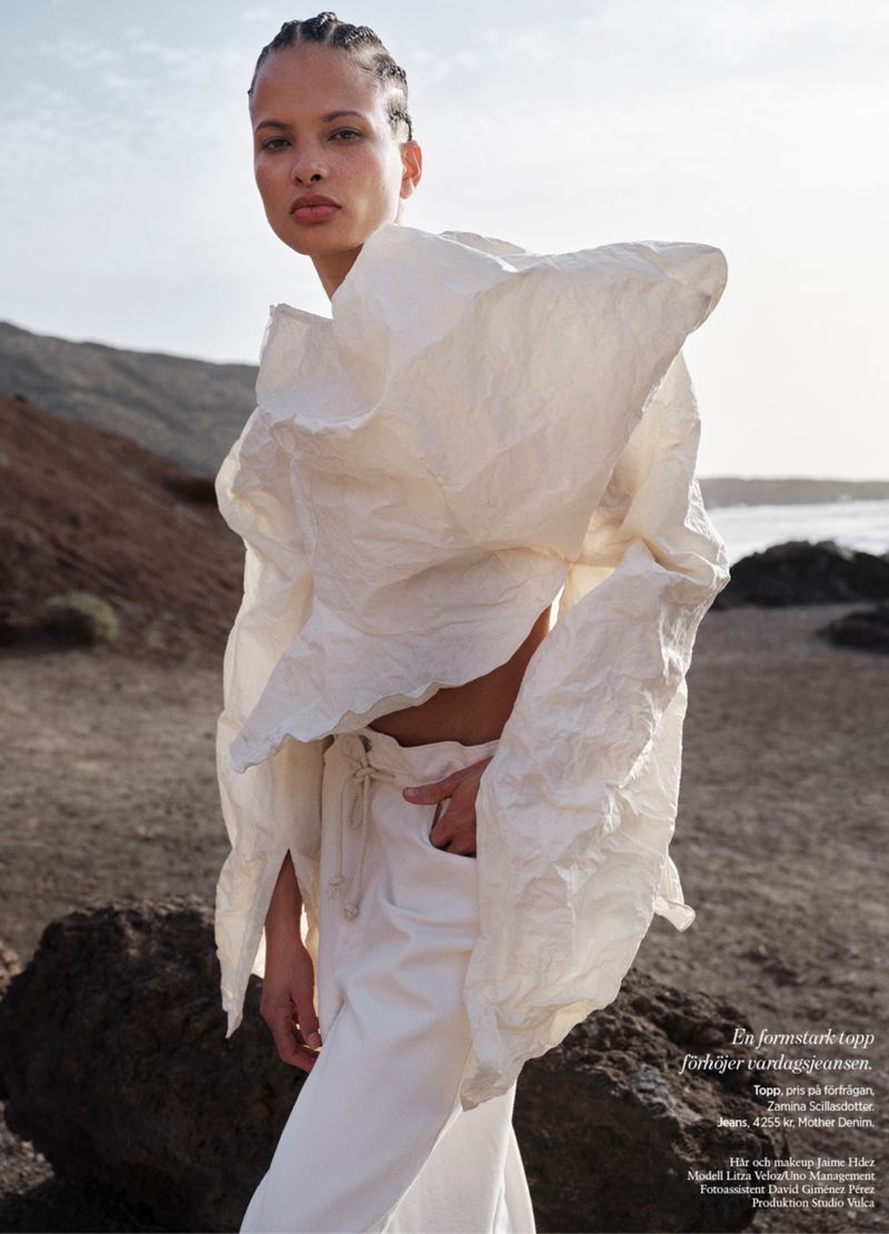 Fashion shot of a Black female model in a white paper-like top and trousers, posing outdoors against a background of mountains and sea.