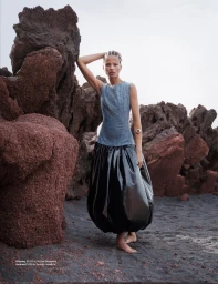 Model leaning against volcanic rock formations wearing a sleeveless grey top and shiny black skirt.