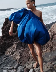 Model wearing a voluminous blue puff dress on a rocky volcanic beach with ocean waves in the background.
