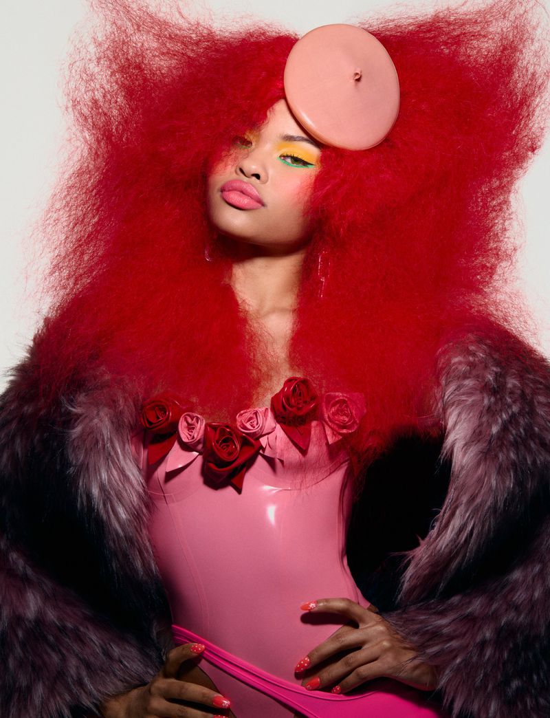 Fashion portrait of a woman with red afro hair, pink latex corset, and fur coat, posing in front of a neutral studio backdrop.