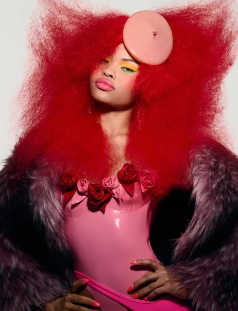 Fashion portrait of a woman with red afro hair, pink latex corset, and fur coat, posing in front of a neutral studio backdrop.