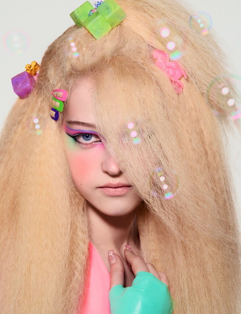 Blonde hair with colorful accessories framing a woman's face with bright makeup, soft pink blush, and blue eyes, against a white background.