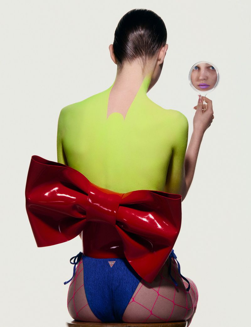 Back view of a female posing with a mirror, neon green body paint, and a large red bow, set against a white backdrop in a studio.
