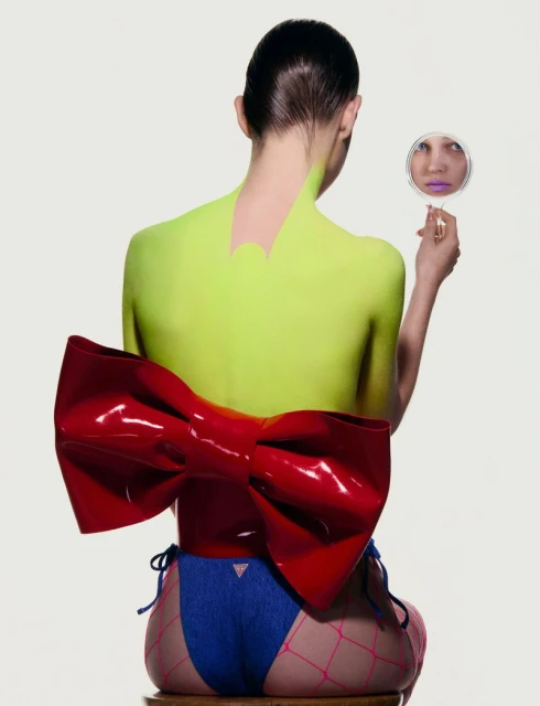 Back view of a female posing with a mirror, neon green body paint, and a large red bow, set against a white backdrop in a studio.