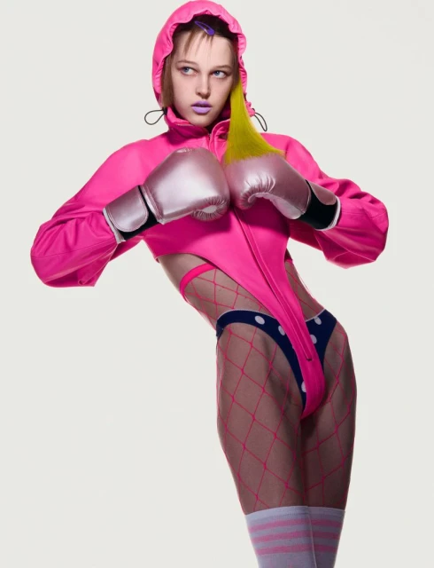 A woman with short hair wears a pink hooded outfit and boxing gloves in a studio shot with a white background and soft lighting.
