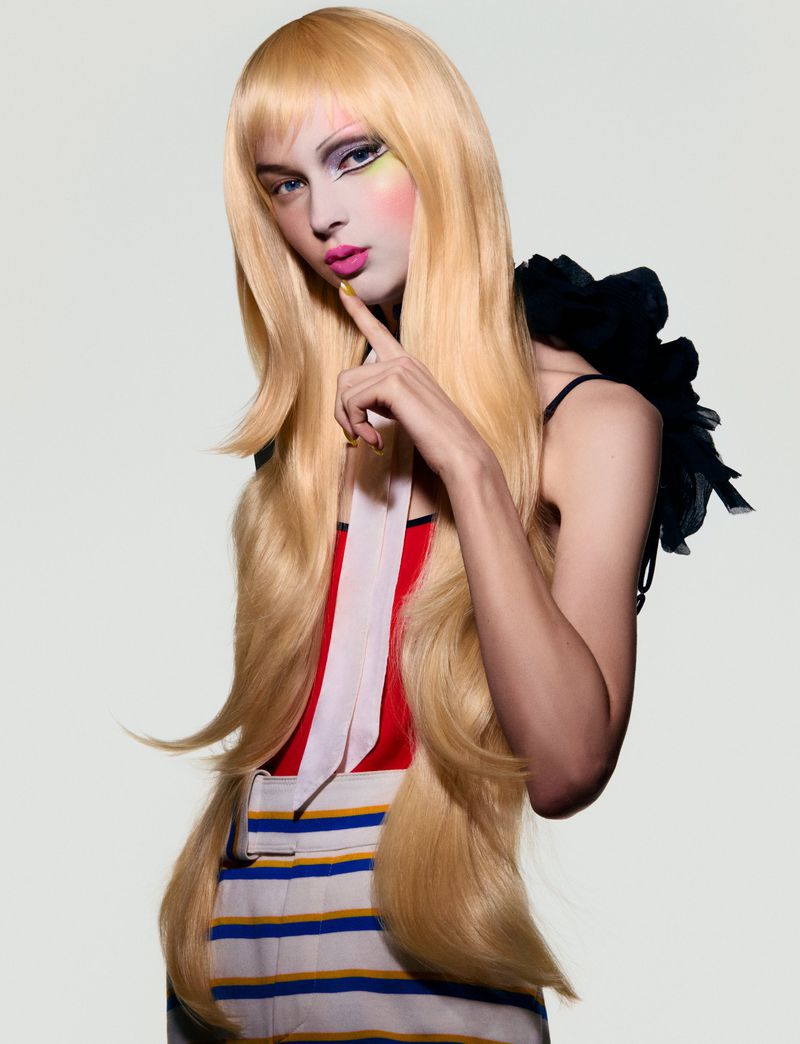 Elegant woman with blonde wig and theatrical makeup poses in a studio setting with black feather accessory and striped dress.