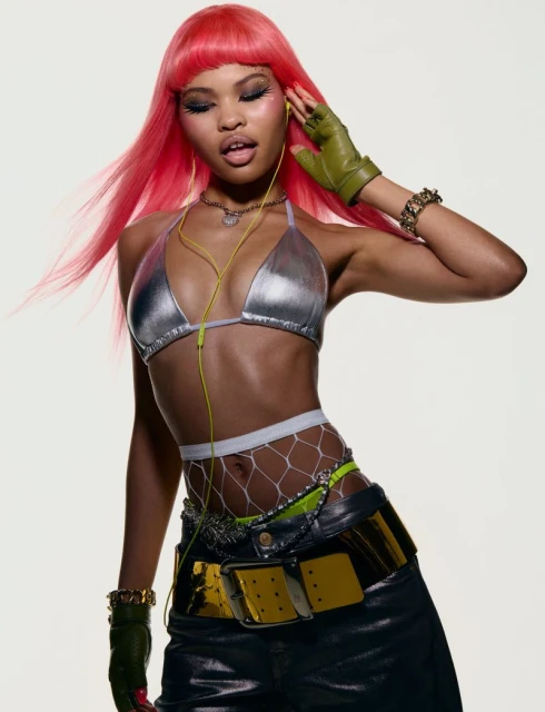 Woman with pink hair poses wearing a silver bikini top, black pants, and wired headphones in a studio setting with a white background.