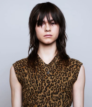Portrait of a model with dark layered hair and fringe wearing a leopard print top against a plain light background.