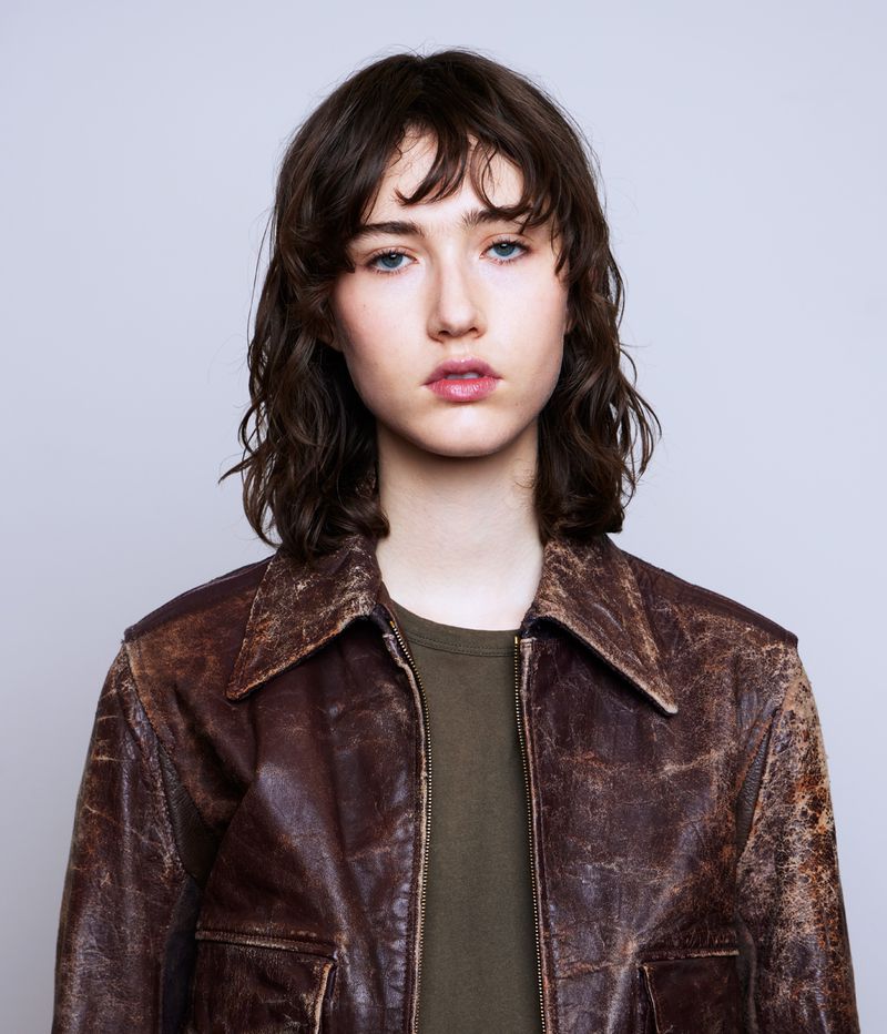 Portrait of a young adult female with shoulder-length wavy brunette hair, wearing a brown leather jacket and an olive-green shirt.
