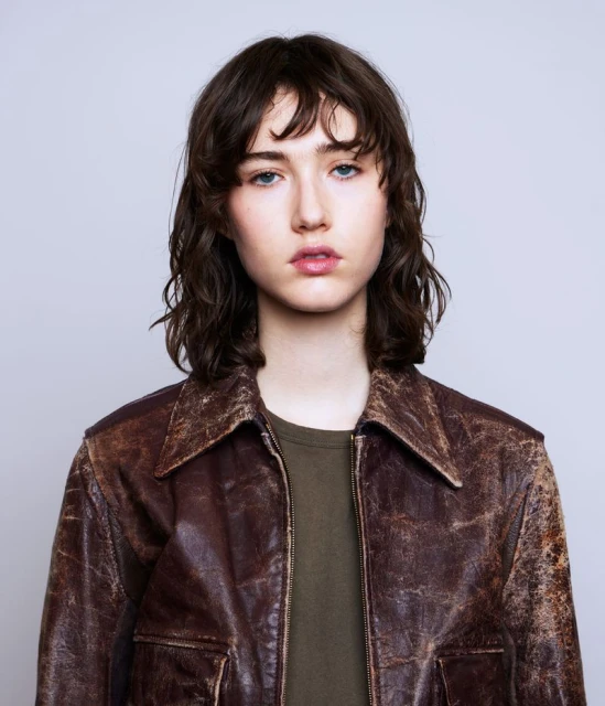 Portrait of a young adult female with shoulder-length wavy brunette hair, wearing a brown leather jacket and an olive-green shirt.