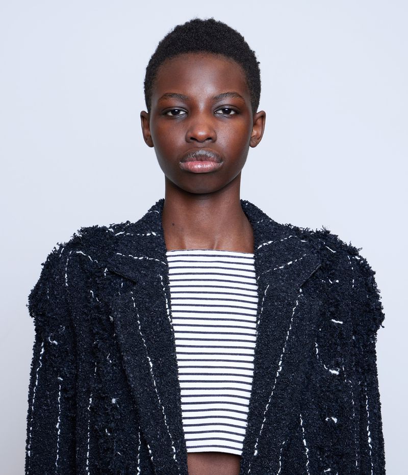 Close-up studio portrait of a young Black woman wearing a striped crop top and a textured blazer, posing against a light gray backdrop.
