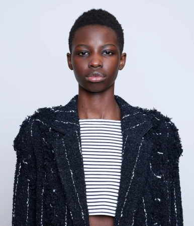 Close-up portrait of model Abuk Manyang Adeer in a dark tweed jacket and striped top against a plain white background.