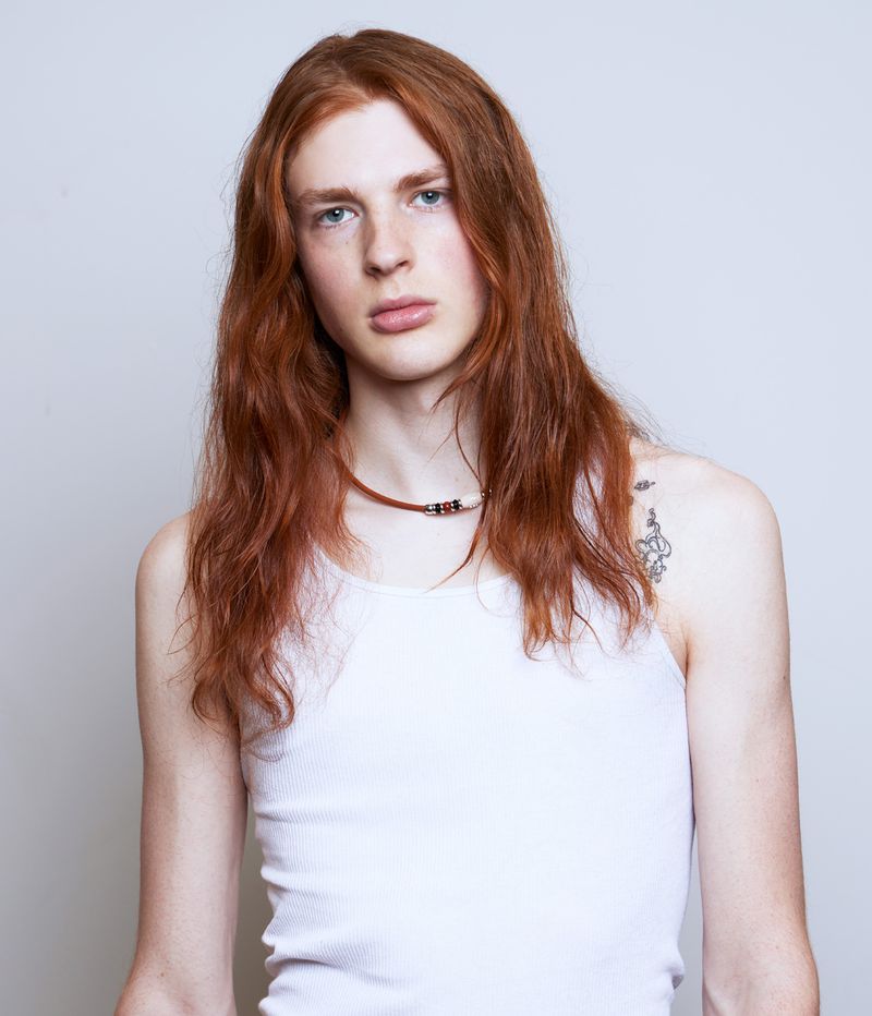 Headshot of a young adult male with long red hair wearing a white ribbed tank top and necklace against a solid grey background.