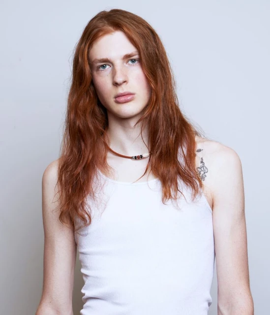 Headshot of a young adult male with long red hair wearing a white ribbed tank top and necklace against a solid grey background.