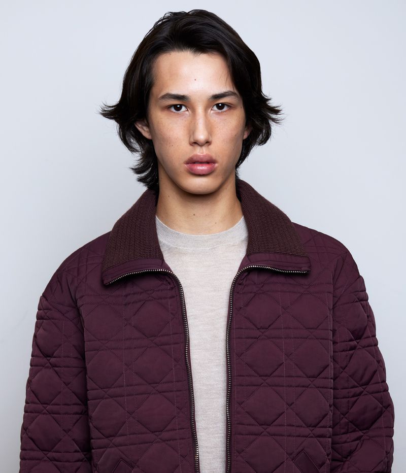 Portrait of a young adult man in a burgundy quilted jacket with a beige shirt, facing the camera in a studio setting.