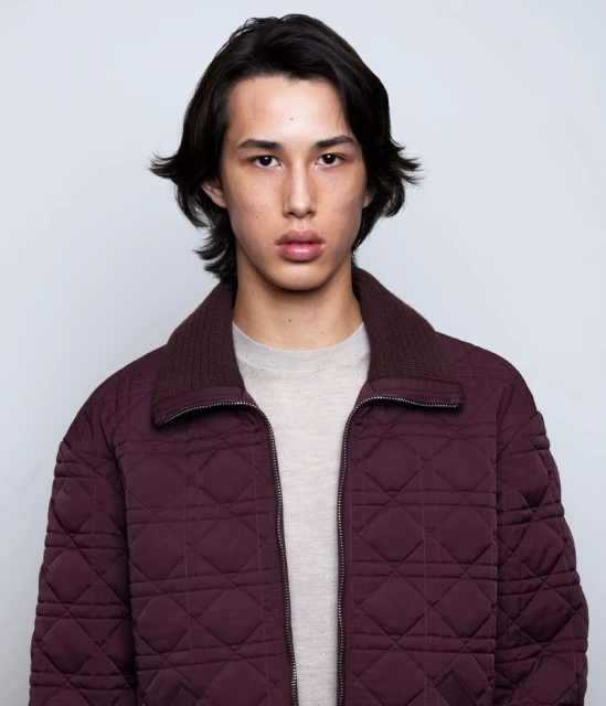 Portrait of a young adult man in a burgundy quilted jacket with a beige shirt, facing the camera in a studio setting.