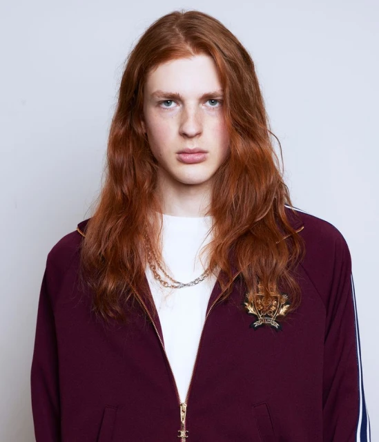 Portrait of a red-haired male fashion model in a burgundy track jacket and gold necklace posing in front of a plain white wall.