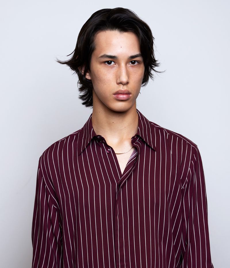 Close-up studio portrait of a young adult male with long dark hair wearing a striped shirt against a white background.