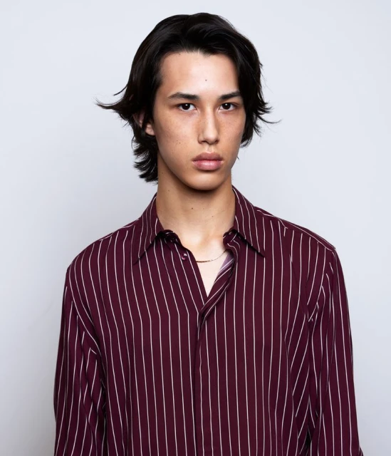 Close-up studio portrait of a young adult male with long dark hair wearing a striped shirt against a white background.