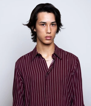 Young man with dark medium-length hair wearing a burgundy striped button-down shirt against a neutral background