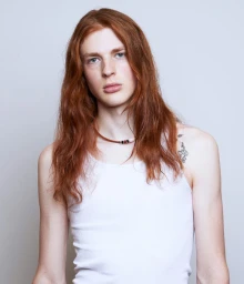 Portrait of young male model with long red hair wearing white tank top against a plain studio background.