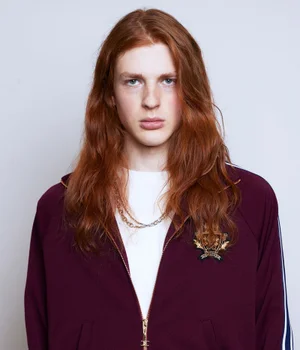 Portrait of a male model with long red hair wearing a burgundy zip-up jacket over a white t-shirt against a plain grey background.