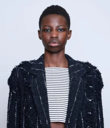 Close-up portrait of model Abuk Manyang Adeer in a dark tweed jacket and striped top against a plain white background.