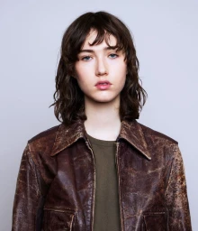 Portrait of a model with dark shoulder-length hair wearing a distressed brown leather jacket against a neutral studio backdrop
