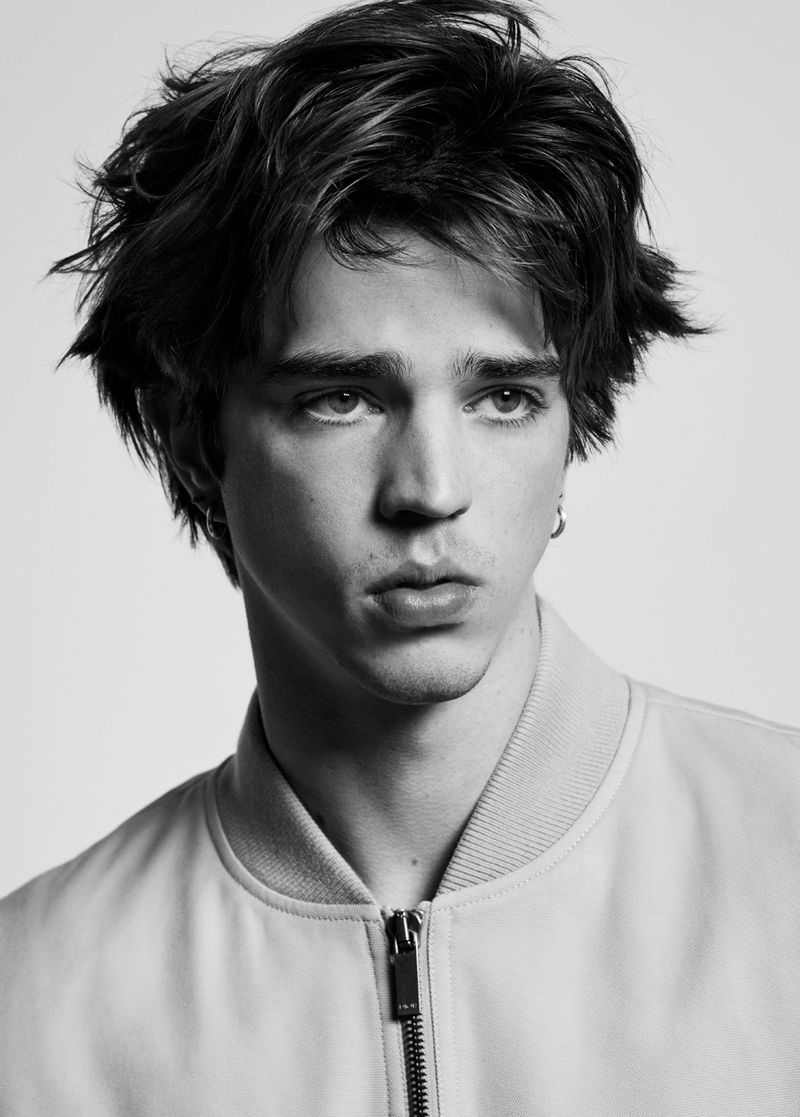 Monochrome studio portrait of a young adult man with tousled brown hair wearing a light-colored bomber jacket, gazing off-camera.