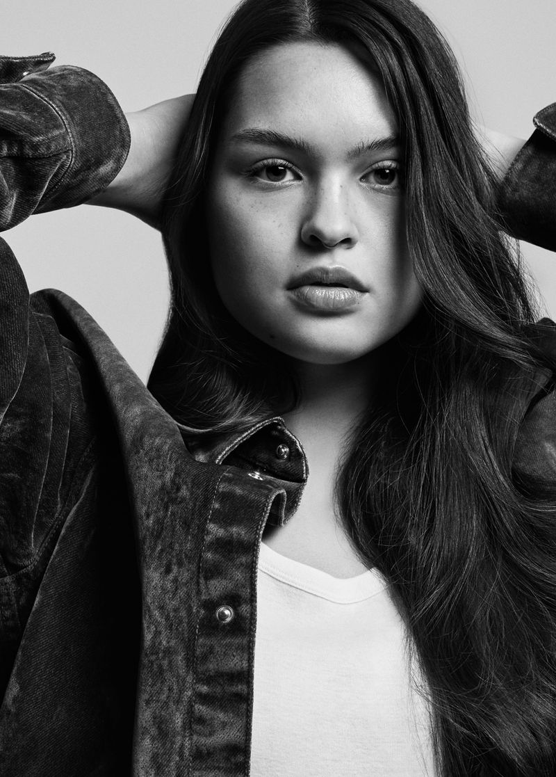 Monochrome portrait of a person with long dark hair posing with hands behind head, wearing a denim jacket over a white t-shirt.