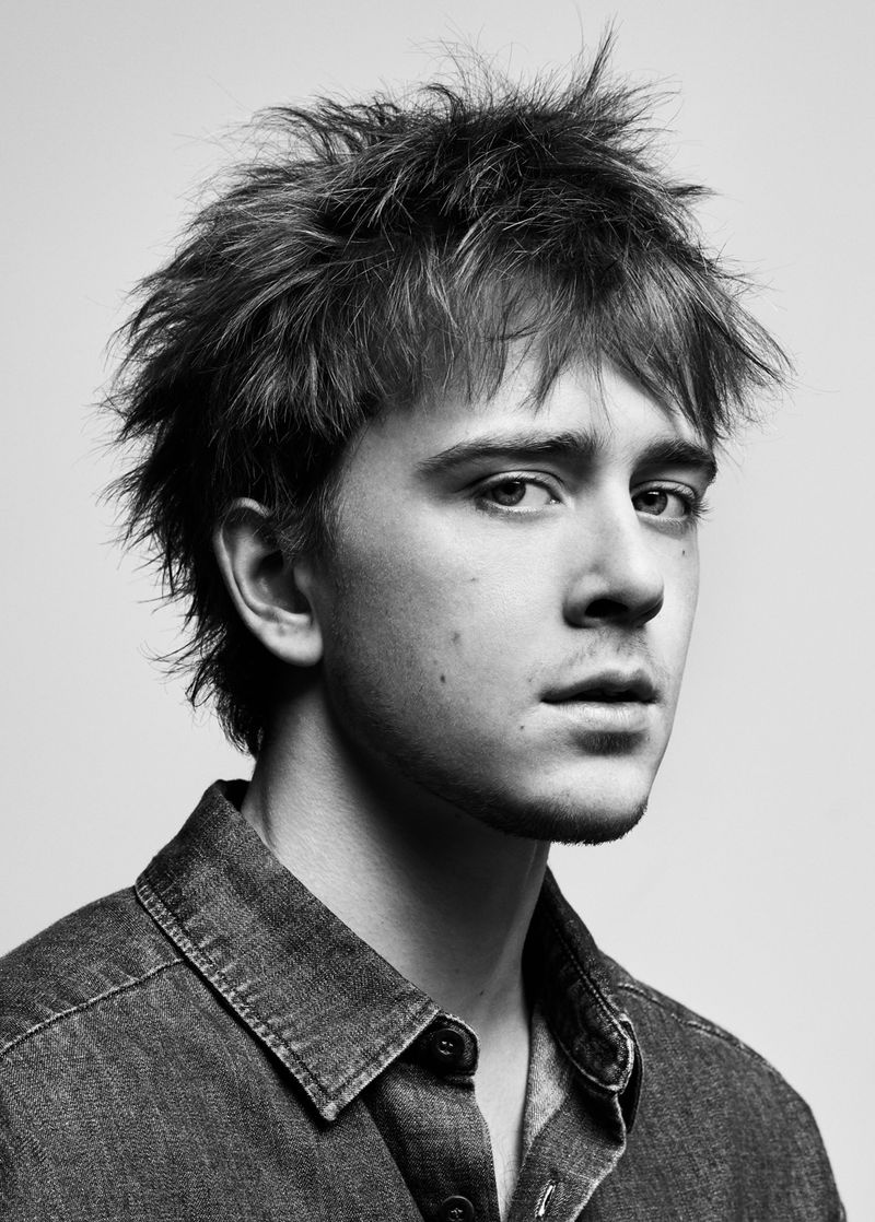 Black and white portrait of a young adult male with dark, spiky hair, wearing a denim shirt, gazing towards the camera in an indoor setting.