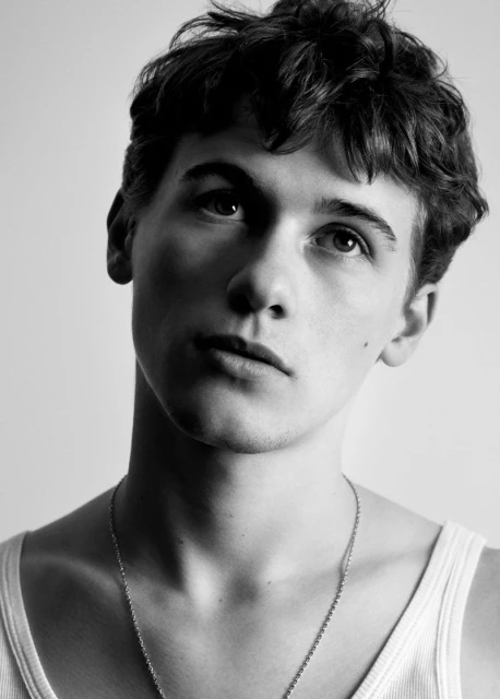 Black and white portrait of a young adult man with dark curly hair, wearing a tank top and a chain necklace, looking up in a studio setting.