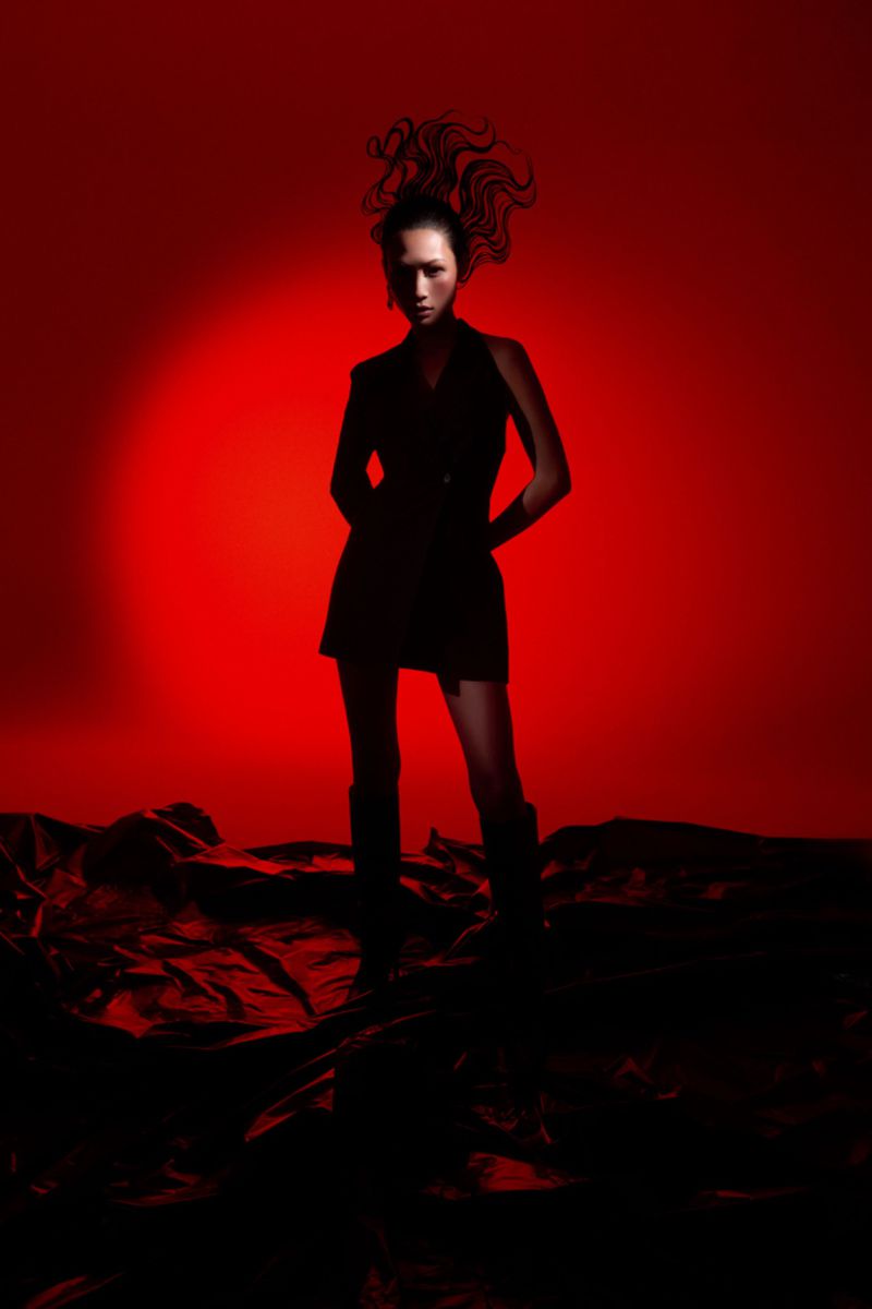 A model poses dramatically in a black dress and boots against a vibrant red backdrop, creating a striking fashion editorial image.