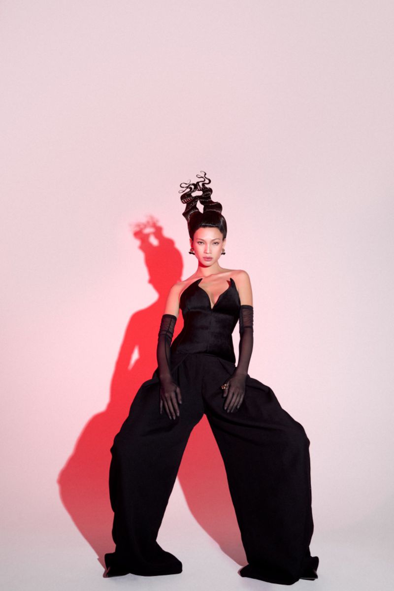 Model in avant-garde black structured outfit and intricate sculptural updo with dramatic red cast shadow on white backdrop